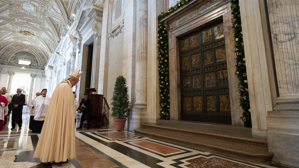 Pope Francis stands before the Holy Door, head bowed in prayer.
